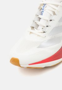 Weißer Sport Schuh mit einem strukturierten Mesh-Obermaterial, silbernen Akzenten, rotem Sohlen-Detail und einer Gummi-Zehenkappe. Schnürverschluss mit flachen Schnürsenkeln.
