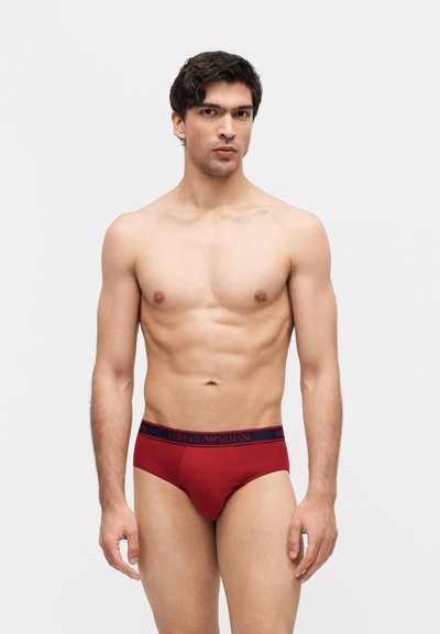 Άνδρας χωρίς πουκάμισο με κοντά σκούρα μαλλιά που φοράει κόκκινα εσώρουχα Emporio Armani με σκούρα μπλε ζώνη μέσης, στέκεται μπροστά σε απλό φωτεινό φόντο.