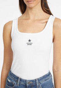 Valge sooniline tops, millel on ruudukujuline kaelus ja keskne tume sinine täht koos "GUESS JEANS" tekstiga all. Sobitatud kõrge taljega teksadega.
