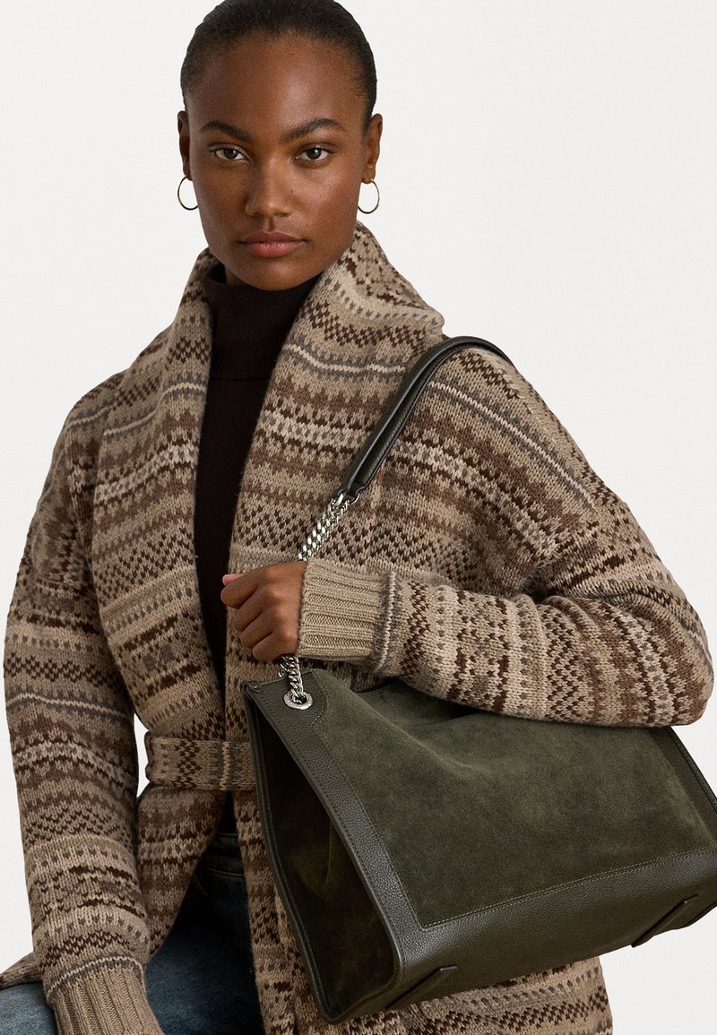 Cardigã castanho com padrão e mangas largas, usado sobre uma blusa escura. O modelo segura uma bolsa de camurça verde com um formato elegante e detalhes em prata.