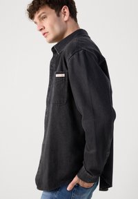 Black denim shirt with a button-down collar and side pocket; features a red and white Calvin Klein label. Loose fit and long sleeves.