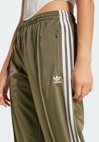 Olivově zelené teplákové kalhoty s bílými bočními pruhy, vyrobené z lesklé látky. Mají elastický pas a kapsu na zip. Zobrazeno logo Adidas.