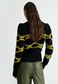 Femme aux cheveux longs et bruns portant un pull noir avec des motifs ondulés rayés jaunes et gris, et un pantalon vert olive, vue de dos.
