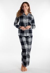 Een glimlachende vrouw staat blootsvoets in een zwart-wit-grijze geruite pyjamastand met knopen tegen een witte achtergrond.