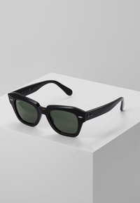 Ochelari de soare pătrați negri cu lentile verzi, cadru gros din plastic, accente argintii pe față și detaliu cu logo pe bradă.