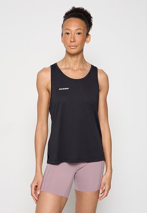 Vrouw staand, gekleed in een zwart mouwloos sporttopje en mauve aansluitende korte broek tegen een effen lichte achtergrond.