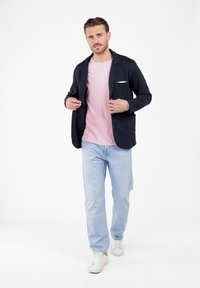 Uomo sistema un blazer blu navy sopra una camicia rosa, indossando jeans azzurri e sneakers bianche, in piedi davanti a uno sfondo bianco.