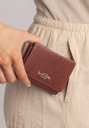 Hand holding brown textured wallet with "Custi Leder studio" logo near beige pants pocket.