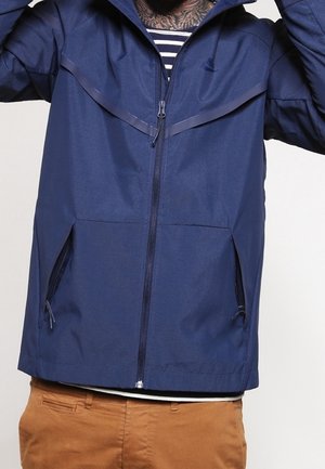 Granatowa kurtka zapinana na zamek z kieszeniami na zamek, noszona na paskowanej koszuli i brązowych spodniach, pokazująca górną część ciała i uniesione ramiona.