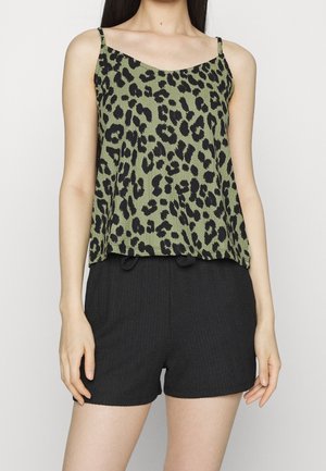 Femme portant un haut vert sans manches avec imprimé léopard noir et un short noir texturé, debout sur fond blanc.