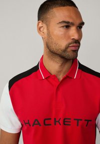 Rotes Poloshirt mit schwarz-weißen Akzenten, kurzen Ärmeln, einem Kragen und dem Logo "HACKETT", das in Schwarz deutlich sichtbar ist.