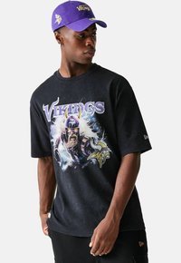 Schwarzes Grafik-T-Shirt mit einem Wikinger-Charakter, lila Akzenten und dem Text "Vikings". Kombiniert mit einer lila Kappe, die das Teamlogo zeigt.