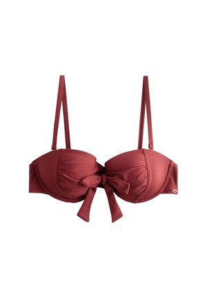 Kobieta z długimi ciemnymi włosami, ubrana w ciemnoczerwony top i dół bikini, stojąca na zewnątrz z zielenią i meblami ogrodowymi w tle.