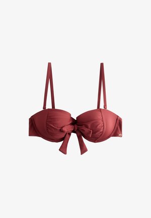 Vrouw met lang donker haar, die een donkerrood bikini top en broekje draagt, staand buiten met groen en tuinmeubilair op de achtergrond.