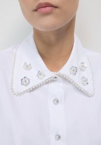 Weißes Hemd mit perlenbesetzten Kanten und dekorativen, durchsichtigen Blumenverzierungen am Kragen, getragen von einer Person.