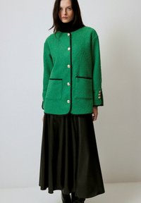 Jachetă verde, texturată, dintr-un amestec de lână cu un design drept, accente negre, detalii cu nasturi aurii și două buzunare frontale, purtată peste o fustă neagră.