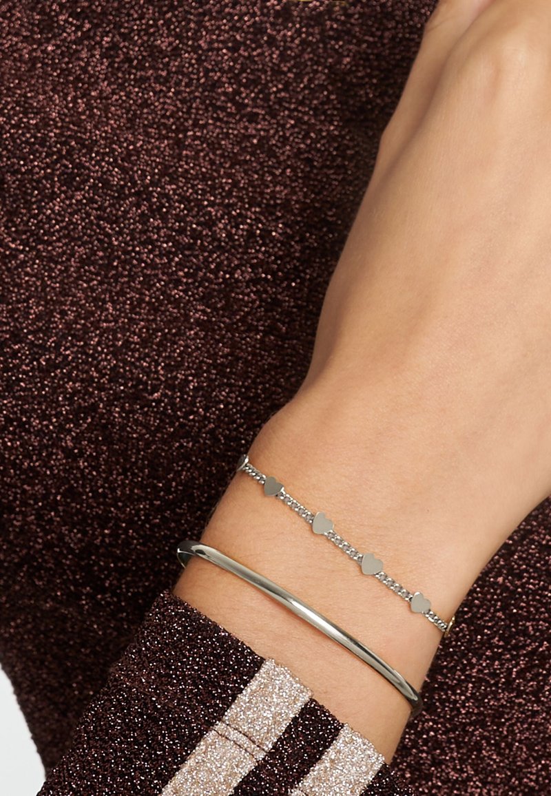 Braccialetti d'argento su un polso: uno è un bracciale rigido in metallo liscio, mentre l'altro presenta piccole forme a cuore alternate a collegamenti lucidi.