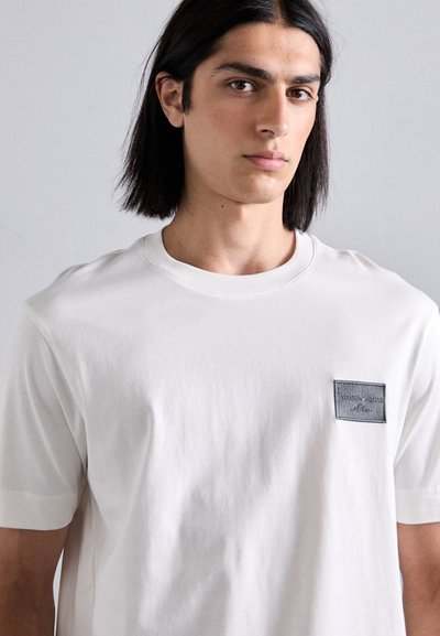 White cotton t-shirt with a crew neck, short sleeves, and a square black patch on the left chest featuring embossed text.