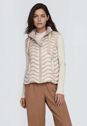 Giovane donna con occhi azzurri che indossa un giubbotto beige trapuntato senza maniche sopra una maglia a maniche lunghe e pantaloni marroni a gamba larga, in piedi davanti a uno sfondo grigio.