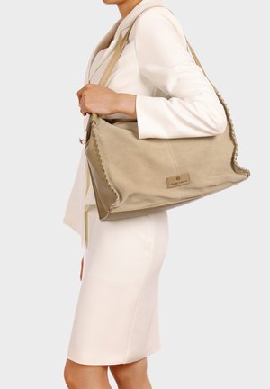 Donna con blazer bianco e gonna che porta una grande borsa beige in pelle scamosciata con bordi intrecciati e tracolle in pelle.