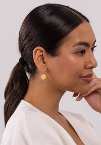 Goldene Creolen mit einem strukturierten runden Anhänger an einem klappbaren Verschluss. Das Model hat lange, dunkle Haare, die zu einem Pferdeschwanz gebunden sind.