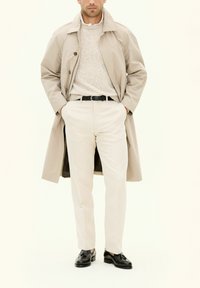 Trench coat beige indossato sopra un maglione chiaro, pantaloni crema e scarpe nere. Caratteristiche notevoli includono bottoni, cintura e texture lisce.