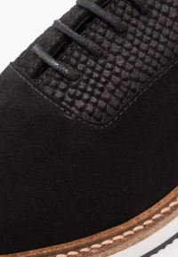 Chaussure noire avec tige texturée à motif écailleux et sections en daim lisse. Lacets noirs, semelle contrastante avec détails de couture visibles.