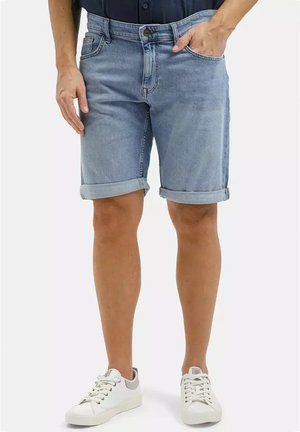 Mężczyzna w jasnoniebieskich, podwiniętych szortach dżinsowych z przednimi kieszeniami i białych sportowych butach, stojący na jednolitym tle.