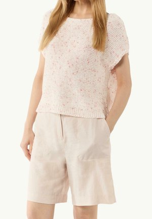 Femme portant un haut rose à manches courtes en maille ample avec des taches et un short taille haute beige clair, une main dans la poche.