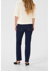 Pull en maille beige clair à manches courtes, associé à un jean bleu foncé à jambe droite et des talons ouverts noirs. Vue de dos.