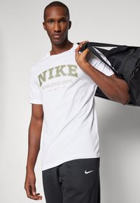 T-shirt bianco in cotone con logo "NIKE" in verde oliva, maniche corte, scollatura rotonda. Il modello tiene una borsa da palestra nera sulla spalla.