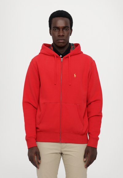 Sudadera roja con cremallera y bolsillo canguro, con un logo dorado en el pecho izquierdo; cuenta con una capucha ajustable y puños acanalados.