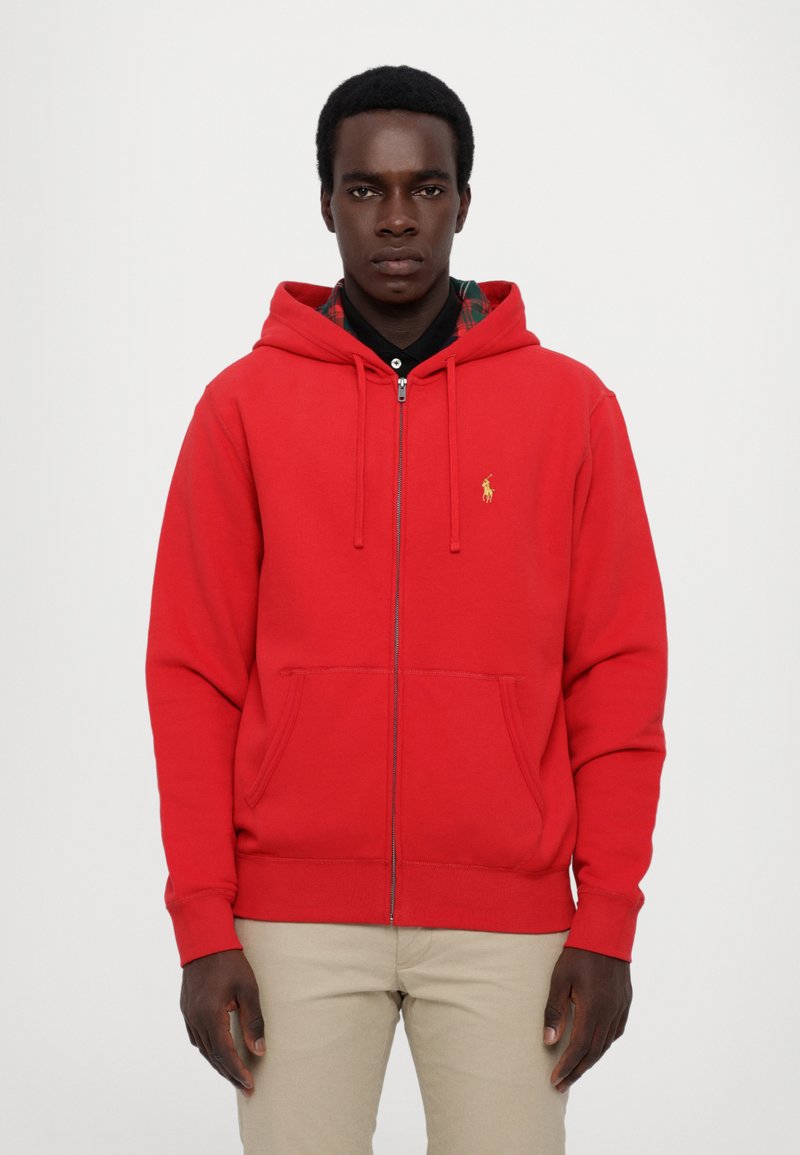 Sudadera roja con cremallera y bolsillo canguro, con un logo dorado en el pecho izquierdo; cuenta con una capucha ajustable y pu ños acanalados.