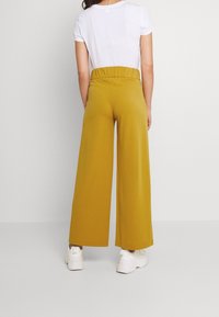 Senfgelbe weitgeschnittene Hose mit elastischem, gerafftem Bund, aus einem glatten Material hergestellt. Tragen mit einem weißen T-Shirt mit kurzen Ärmeln.