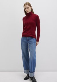 Roter Rollkragenpullover mit Kabelstricktextur, kombiniert mit blauen Jeans mit weitem Bein und aufgerollten Säumen sowie schwarzen Knöchelstiefeln.