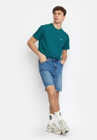 Teal katoenen t-shirt met korte mouwen en een klein logo, gecombineerd met blauwe denim shorts en beige sneakers met witte sokken.