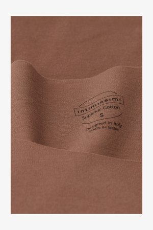 Bomullstyg i varm brun nyans med en mjuk yta. Har en svart tryckt etikett som anger märke, storlek och produktionsdetaljer.