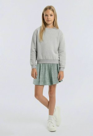 Noor tüdruk seisab ristatud jalgadega, kandes halli dressipluusi, mustrilist seelikut, valgeid sokke ja valgeid tossusid tavalise tausta ees.