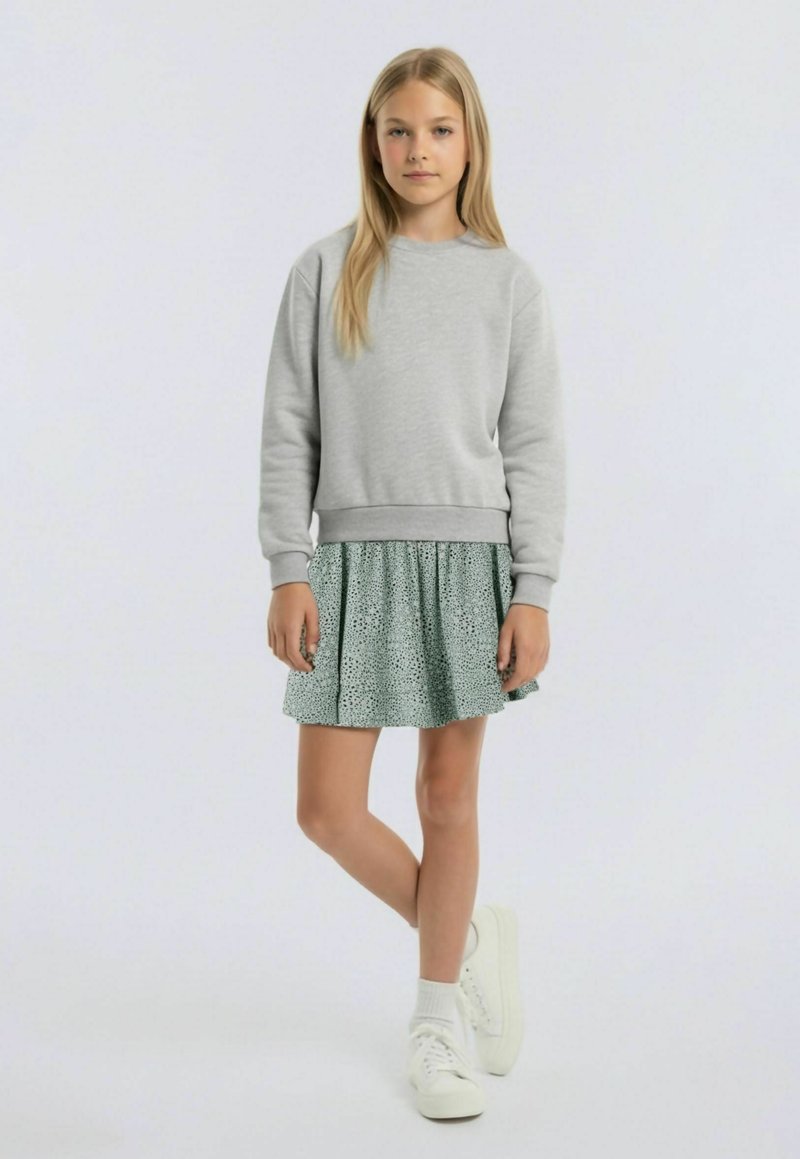 Jeune fille debout, jambes croisées, portant un sweat-shirt gris, une jupe à motifs, des chaussettes blanches et des baskets blanches sur un fond uni.