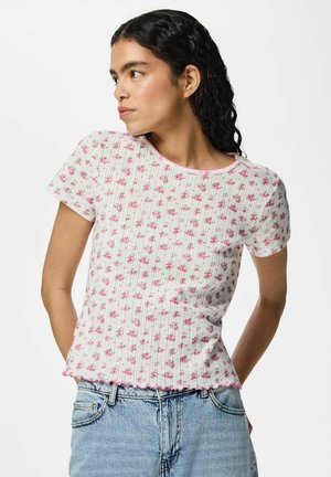 Ung kvinde med krøllet mørkt hår iført hvid ribbet kortærmet top med små lyserøde blomster og lyseblå jeans, kigger mod venstre.