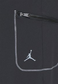Los pantalones deportivos negros cuentan con un bolsillo con cremallera, costuras grises y un logo "Jumpman" blanco. Tienen una textura suave y un diseño ajustado.