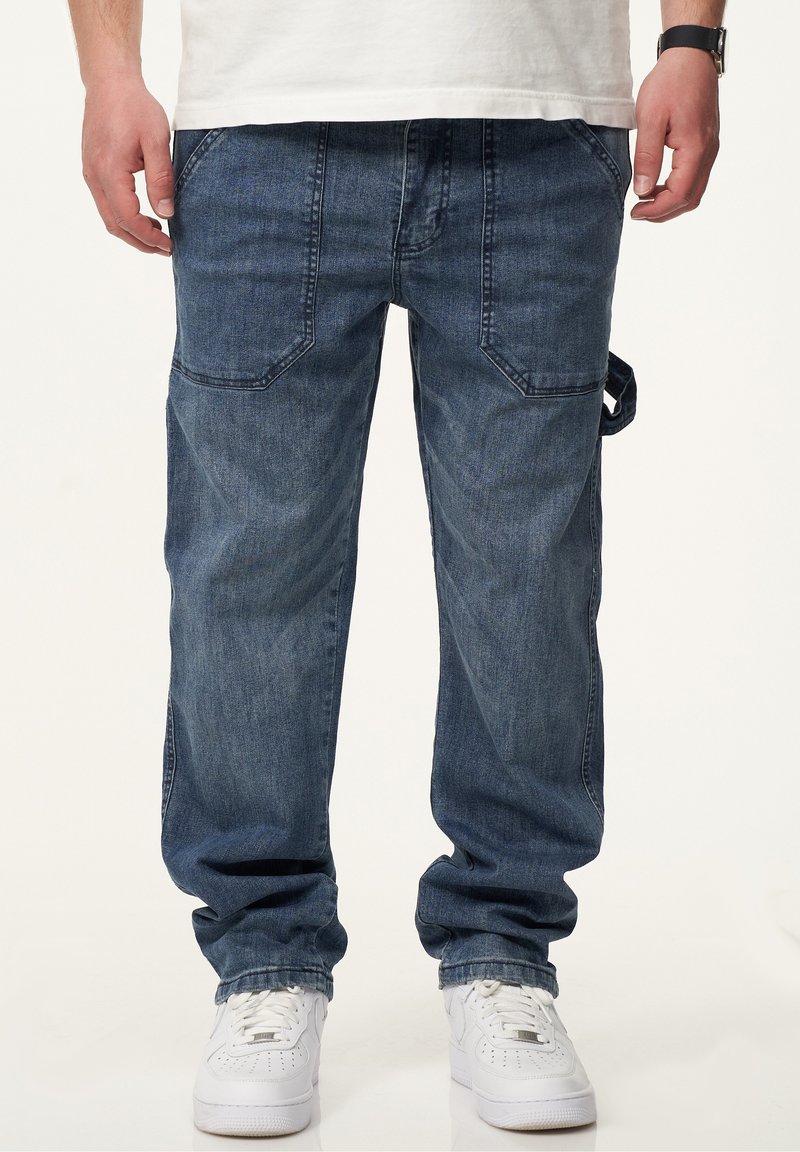 Dunkelblaue Denim-Cargohosen mit lockerer Passform, ausgefallenen Taschenformen und seitlichem Schlaufen-Detail, kombiniert mit weißen Sneakern.