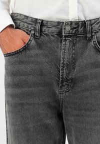 Man met vervaagde zwarte denim jeans, hand in de voorkzak en een wit shirt dat is ingestopt.