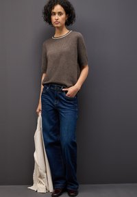 Maglione a maglia marrone con maniche corte, collo crema e jeans a gamba larga blu. Tiene in mano una giacca beige, in piedi contro uno sfondo grigio.