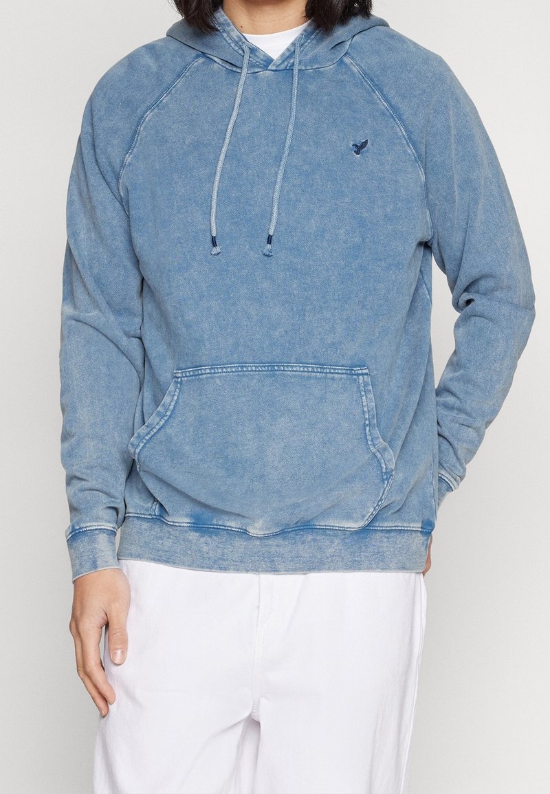 Blekt blå hoodie med kängurufickor, dragsko i huvan och ribbade ärmslut. Har en subtil logotyp på bröstet. Mjuk, texturerad tyg.