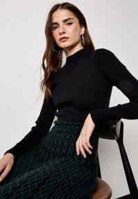 Donna con capelli castani lunghi che indossa orecchini a cerchio d'argento, maglione nero a coste e gonna verde a motivi, seduta su una sedia contro una parete liscia.