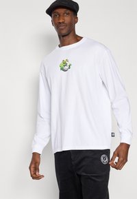 Λευκό βαμβακερό μακρυμάνικο μπλουζάκι με πράσινο σχέδιο αρκούδας στο στήθος, συνδυασμένο με μαύρο παντελόνι από κορντόνι και μαύρο καπέλο.