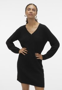 Robe tricotée noire à col en V, à manches longues, en tissu texturé, avec un détail de bouton sur le côté. Coupe drapée atteignant le haut des cuisses. Design simple et polyvalent.