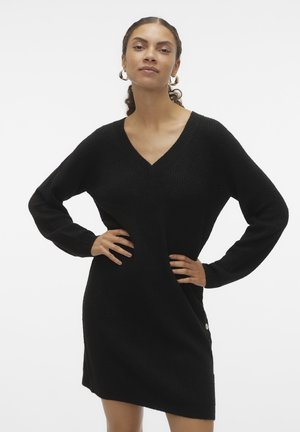 Robe tricotée noire à col en V, à manches longues, en tissu texturé, avec un détail de bouton sur le côté. Coupe drapée atteignant le haut des cuisses. Design simple et polyvalent.