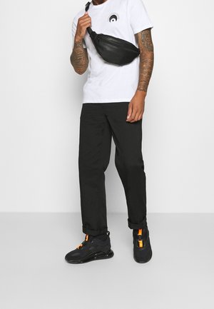 Schwarze Leder-Bauchtasche, diagonal getragen, weißes T-Shirt mit kreisförmigem Motiv, schwarze Hose und schwarze Turnschuhe mit orangefarbenen Akzenten.